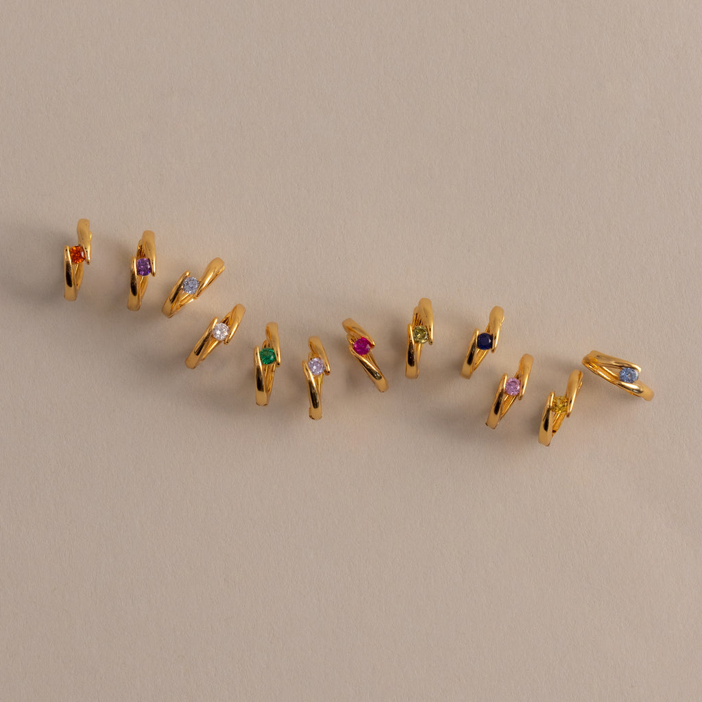 Floating Birthstone Huggies—gold hoops with vibrant gemstones—are arranged diagonally on a beige surface, ideal for fans of personalized jewelry.