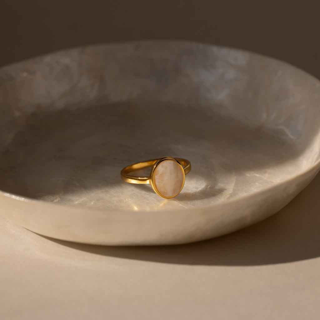 The Moonstone Signet Ring, featuring a round, light moonstone, rests in a cream ceramic dish—a refined and elegant accessory for daily wear.