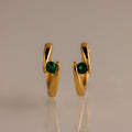 Floating Emerald Huggies, featuring gold hoops with green emerald-like stones, stand upright on a smooth, neutral background.