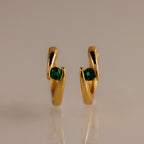 Floating Emerald Huggies, featuring gold hoops with green emerald-like stones, stand upright on a smooth, neutral background.