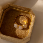 The Cosmic Moonstone Ring, featuring three white stones, shines in a textured beige jewelry box under soft natural light—a stunning choice for boho jewelry lovers.