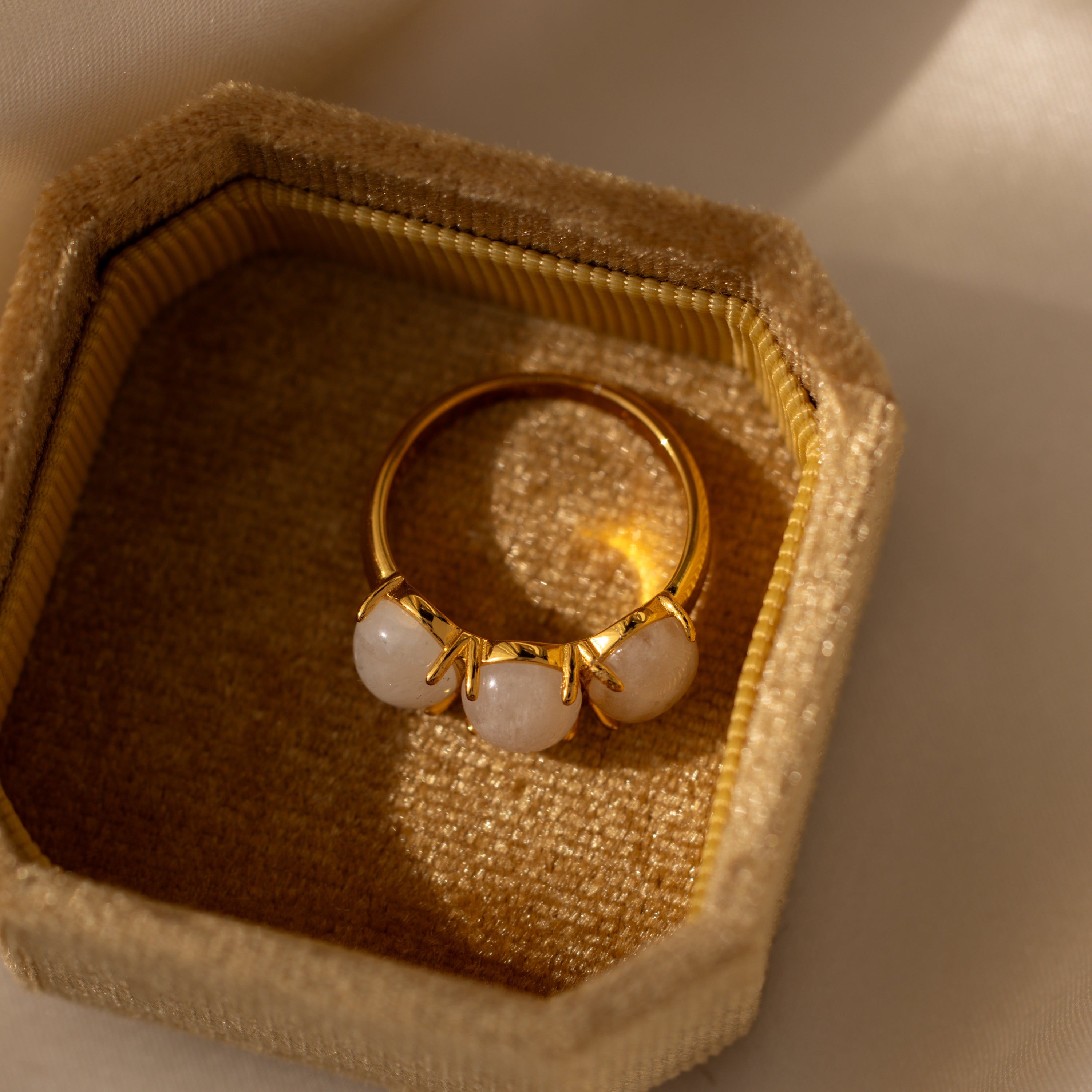The Cosmic Moonstone Ring, featuring three white stones, shines in a textured beige jewelry box under soft natural light—a stunning choice for boho jewelry lovers.