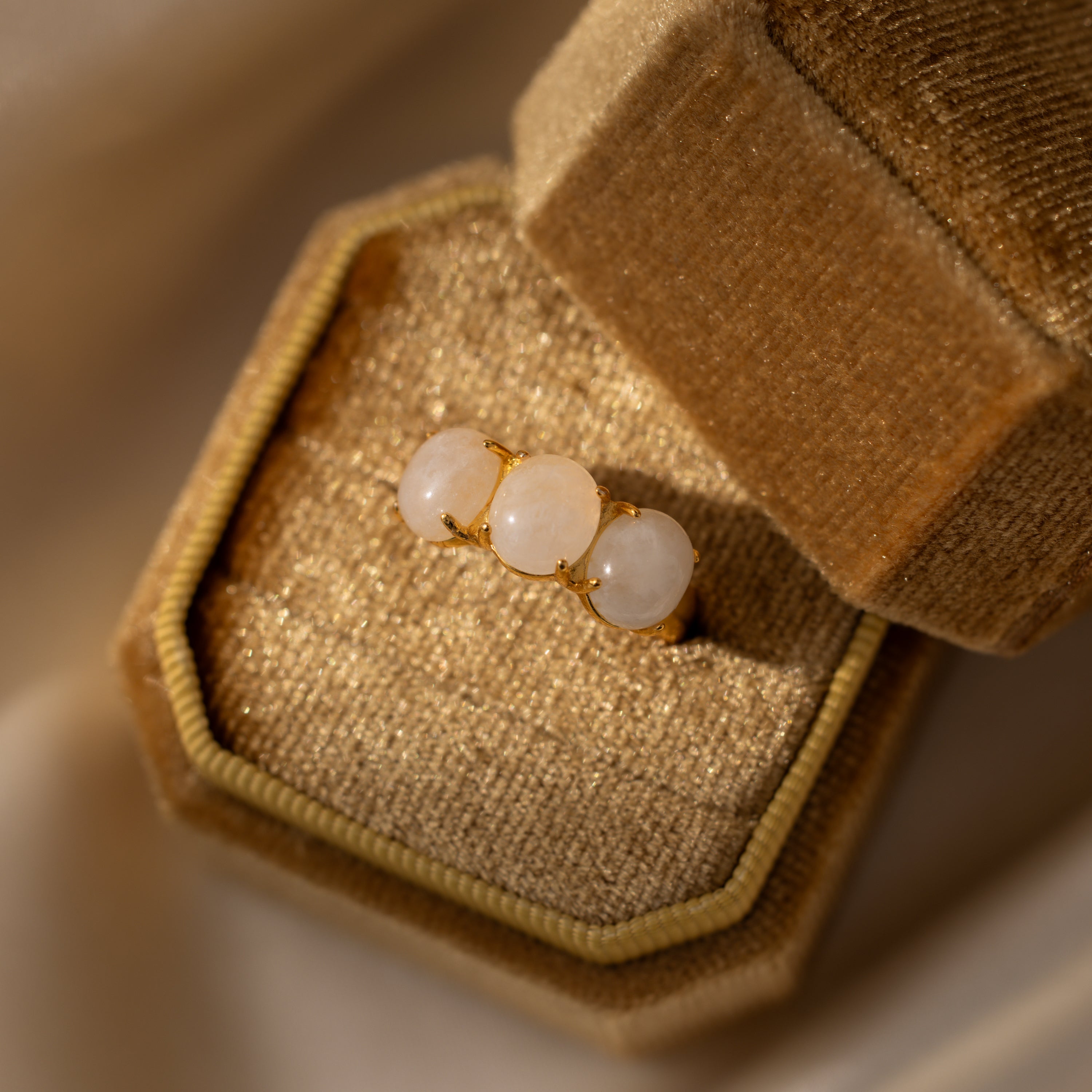 The Cosmic Moonstone Ring—a gold statement piece with three round white stones—comes in a beige velvet ring box, ideal for boho jewelry lovers.