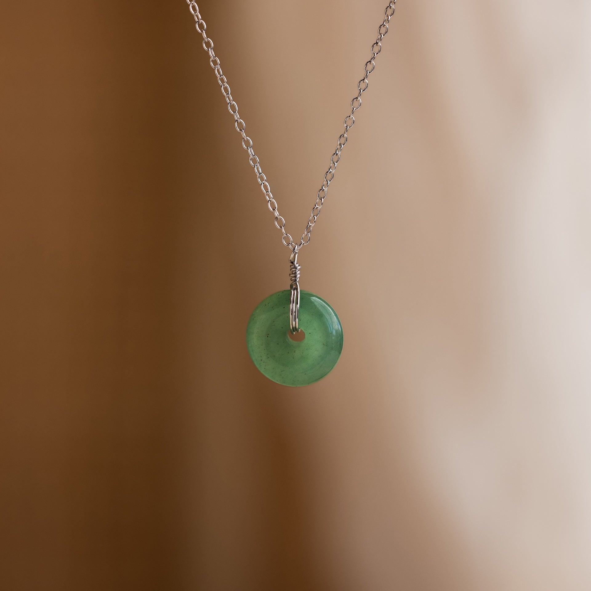 The Round Jade Pendant Necklace features a silver chain and jade gemstone, offering natural elegance against a soft beige background.