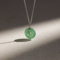 The Round Jade Pendant Necklace in Silver features a jade gemstone that exudes natural elegance, with its green pendant casting a soft shadow on any surface.