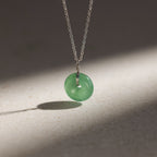 The Round Jade Pendant Necklace in Silver features a jade gemstone that exudes natural elegance, with its green pendant casting a soft shadow on any surface.