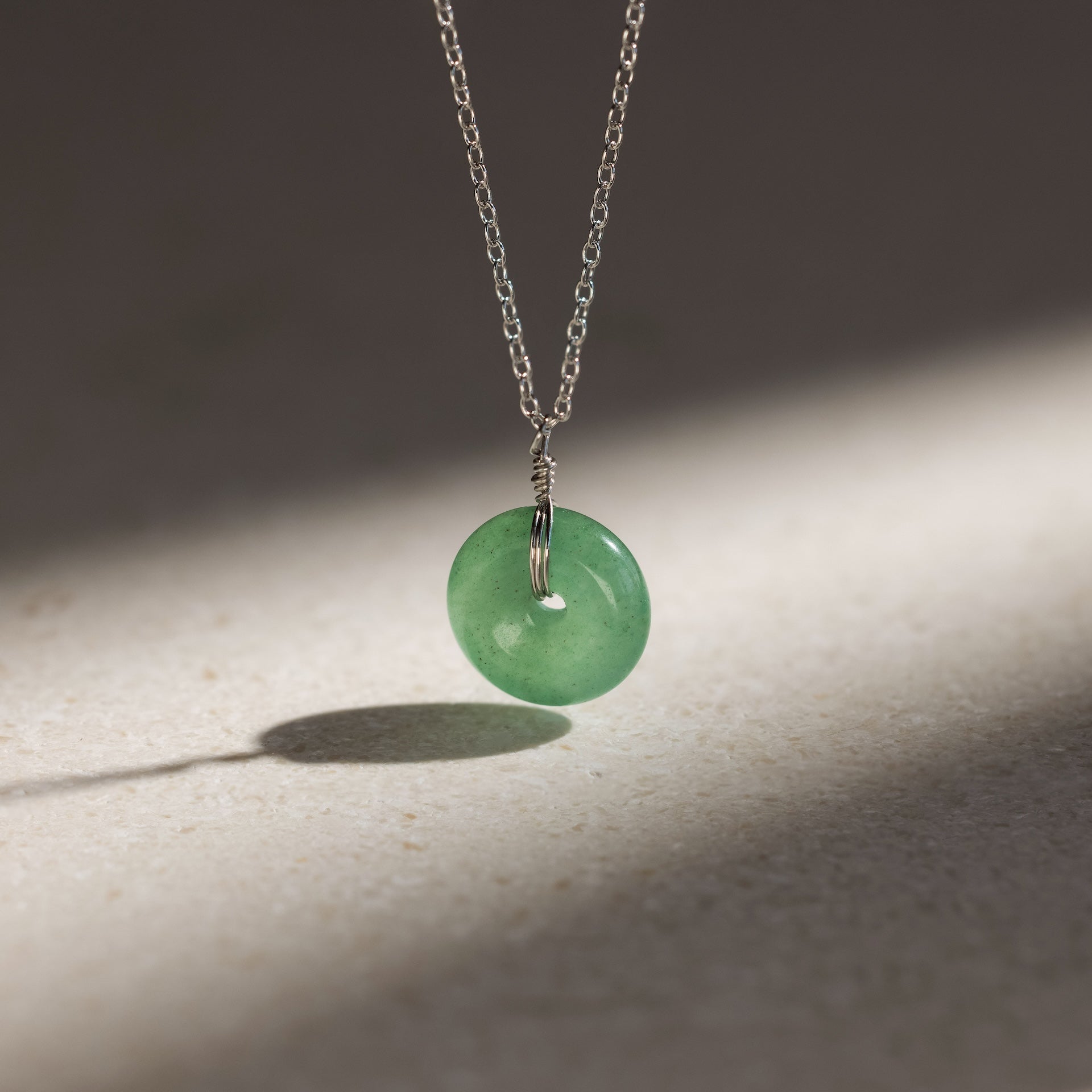 The Round Jade Pendant Necklace in Silver features a jade gemstone that exudes natural elegance, with its green pendant casting a soft shadow on any surface.