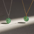 A Round Jade Pendant Necklace in 18K Gold casts an elegant shadow as its jade gemstone hangs gracefully from a gold chain on a light surface.