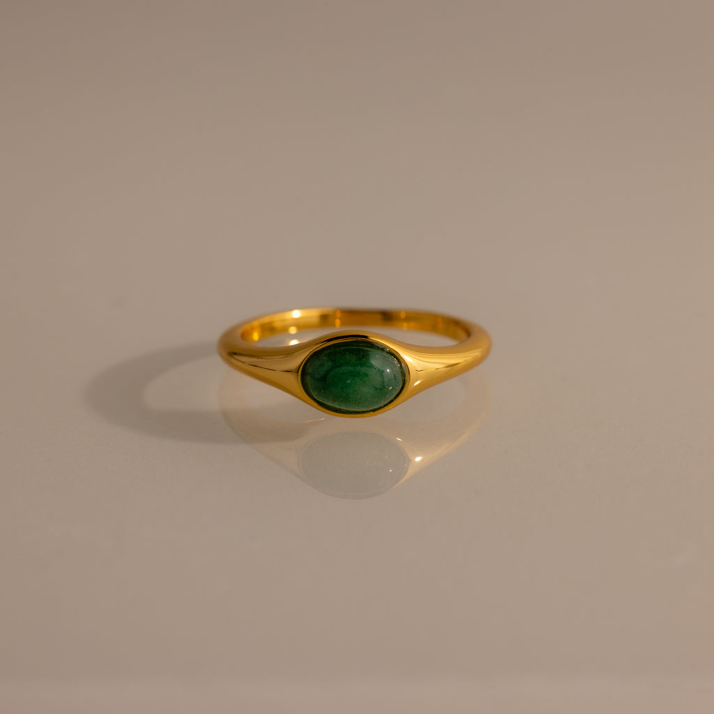 The Oval Jade Signet Ring showcases a green jade stone set in gold, elegantly presented on a reflective surface.