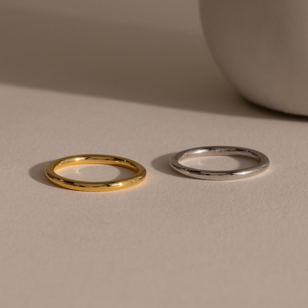 The Minimal Stacking Ring in gold and silver rests side by side on a beige surface, with a white round object nearby.