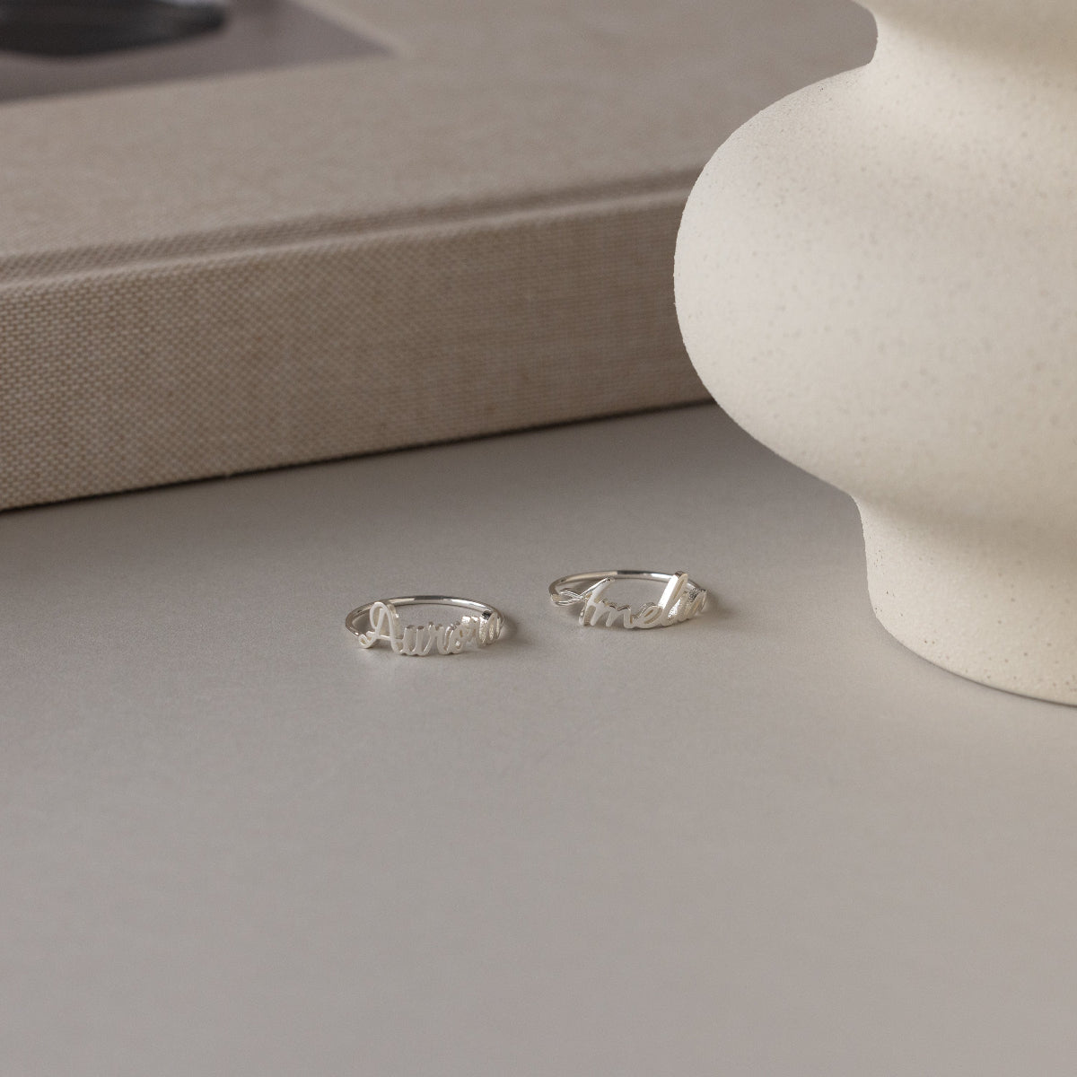 Two sterling silver rings with elegant cursive rest on a light surface by a textured vase and beige book—ideal as personalized rings like the Grace Script Name Ring in Sterling Silver, crafted in an exquisite script font.