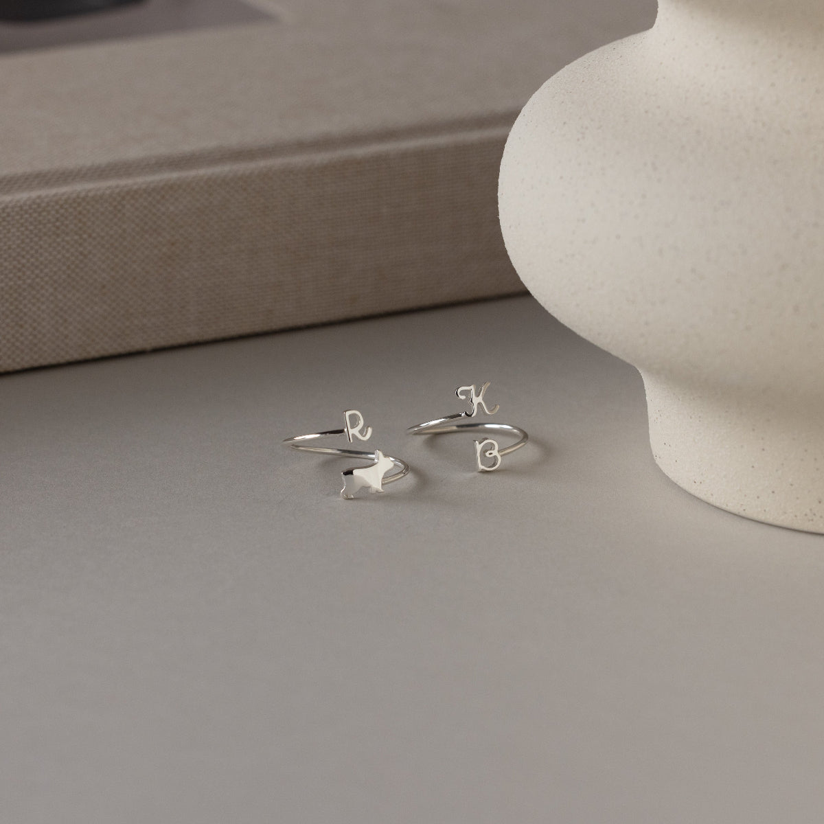 Displayed near a vase and book, the Duo Pet Initial Wrap Ring in Sterling Silver features elegant letter charms—making it the perfect personalized pet ring for any animal lover.