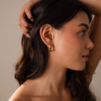 A woman with long dark hair wears Spiral Swirl Hoops, gazing to the side with her hand on her head—an ideal accessory for boho vacation outfits.