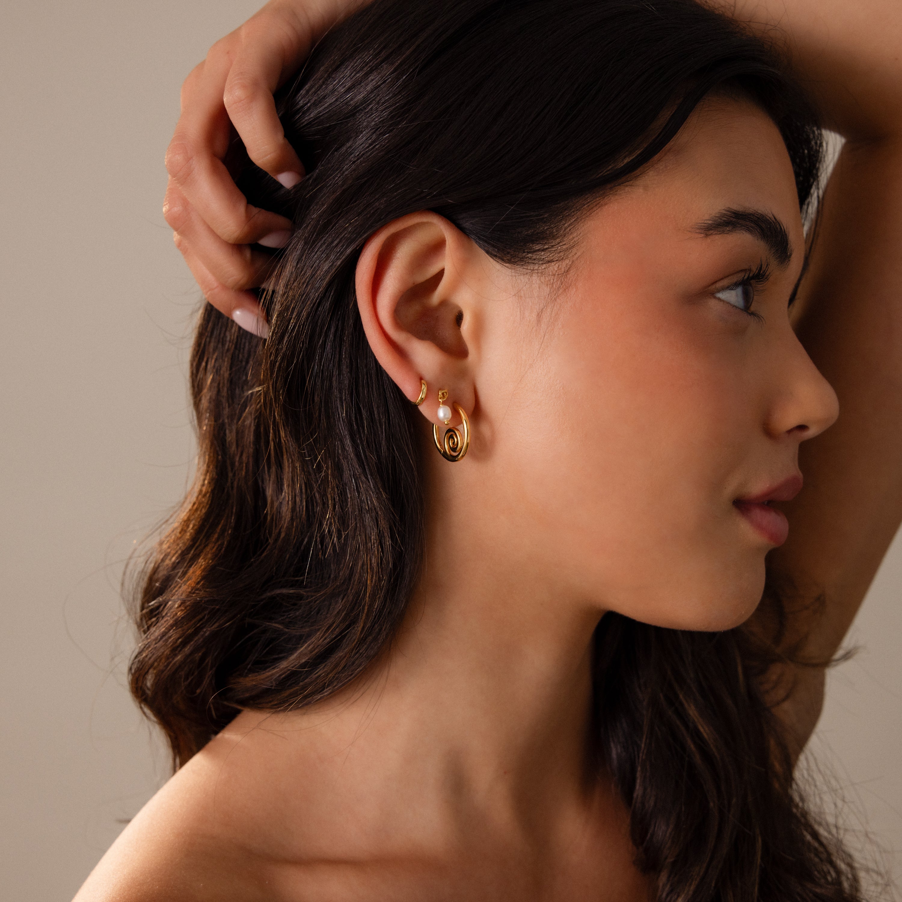A woman with long dark hair wears Spiral Swirl Hoops, gazing to the side with her hand on her head—an ideal accessory for boho vacation outfits.