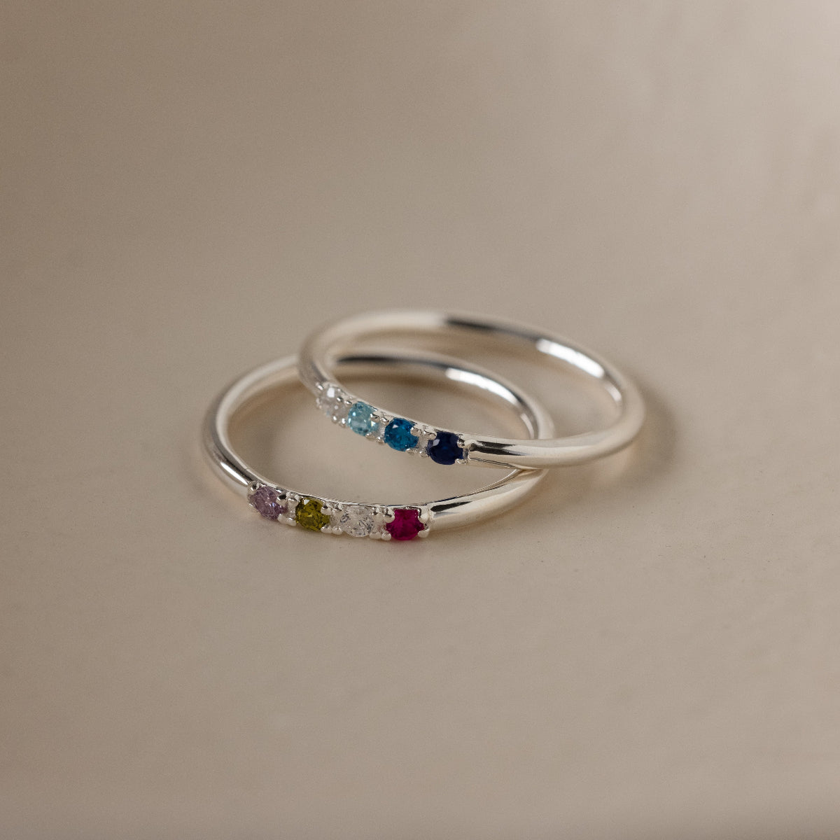 The Mini Birthstone Ring in Sterling Silver features colorful gemstones set in a row—ideal as a dainty accessory or personalized birthstone ring for any occasion, displayed on a smooth beige surface.