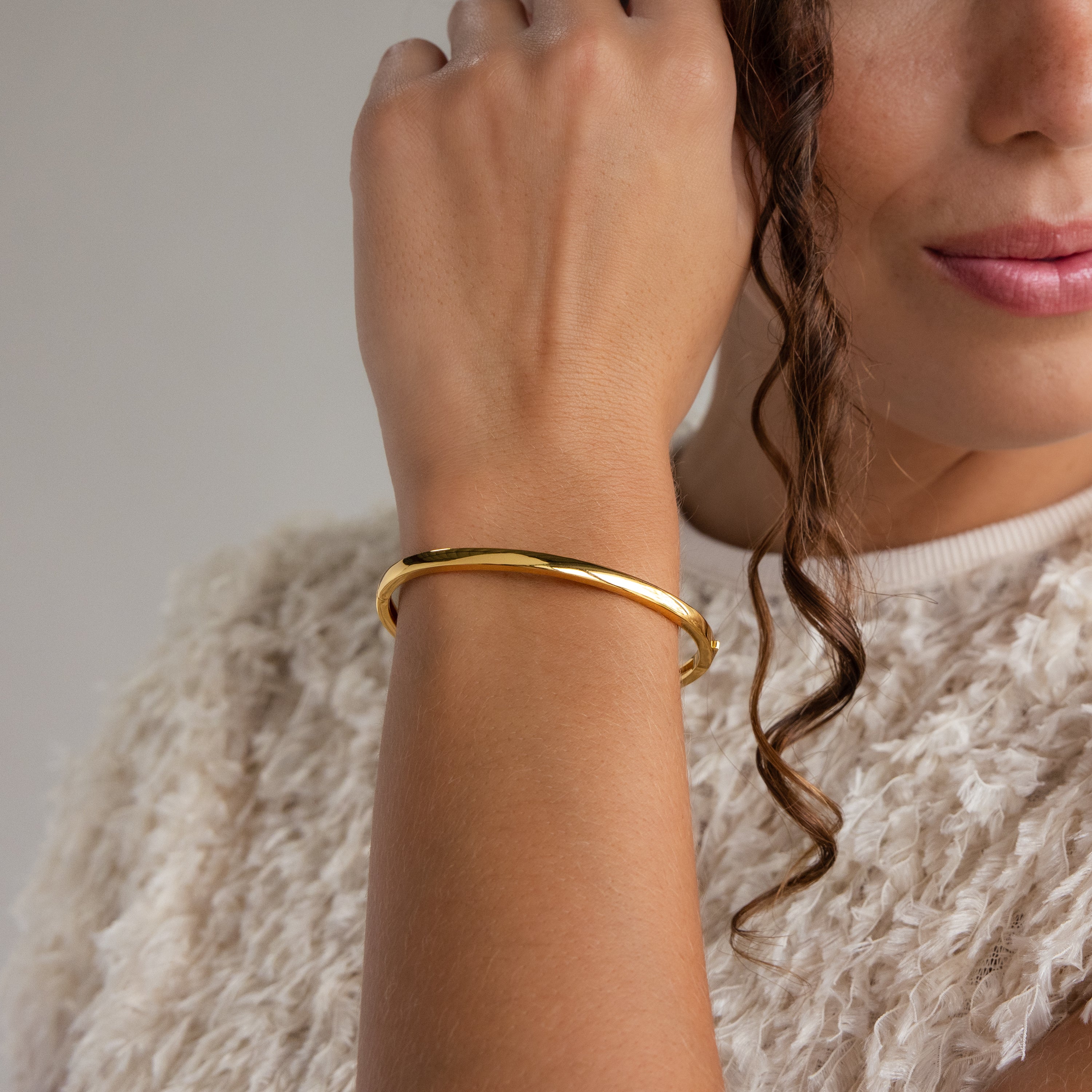 A woman with curly hair and a textured cream top poses with her hand near her face, showcasing the Classic Bangle in 18K Gold—a perfect example of minimalist jewelry style.