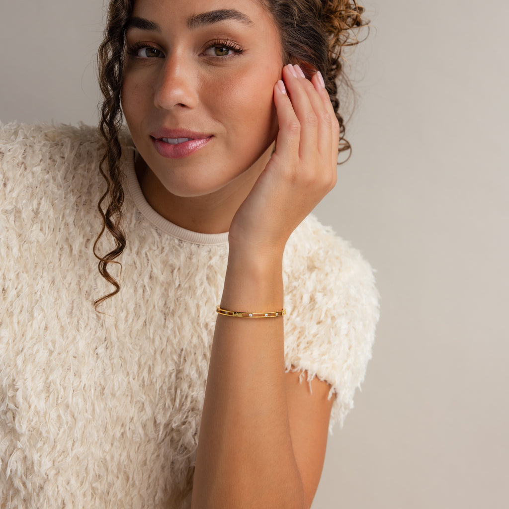 A woman with curly hair smiles softly, touching her face while wearing a white textured top and the elegant Lace Diamond Bangle.