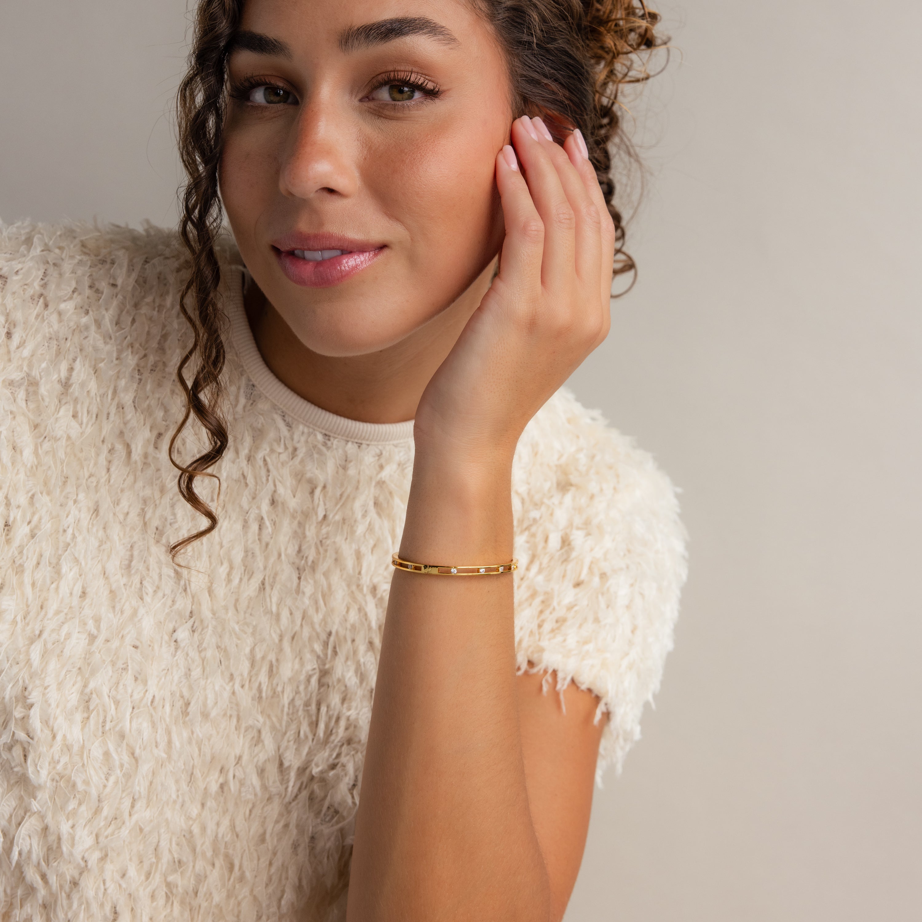 A woman with curly hair smiles softly, touching her face while wearing a white textured top and the elegant Lace Diamond Bangle.