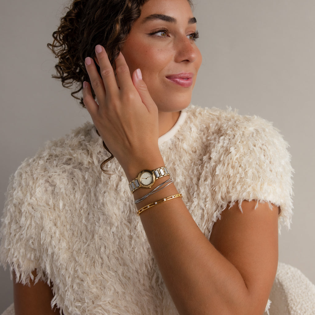 A woman in a textured white top smiles and looks off to the side, posing with her gold bracelets, including the Lace Diamond Bangle in Sterling Silver with round diamonds and an open station design, next to her watch.