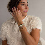 A woman in a textured white top smiles and looks off to the side, posing with her gold bracelets, including the Lace Diamond Bangle in Sterling Silver with round diamonds and an open station design, next to her watch.