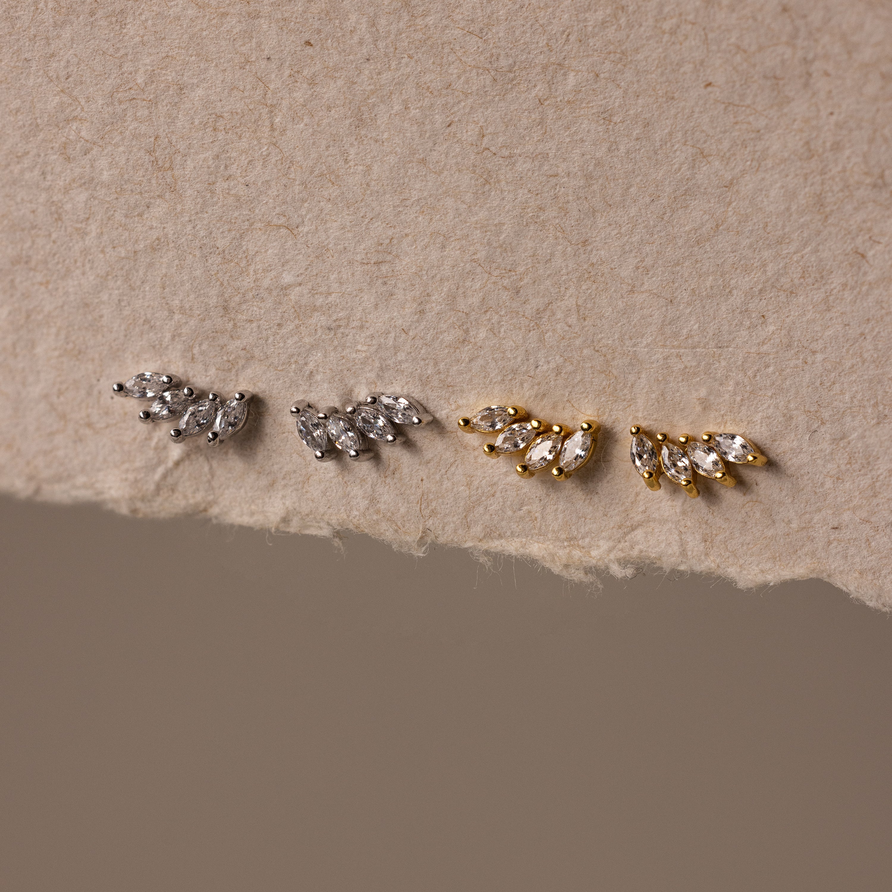 Two pairs of Marquise Diamond Climbers earrings—one silver, one gold—in a leaf-shaped design on textured beige paper; ideal wedding jewelry accents.