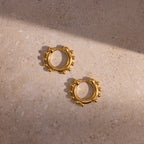 A pair of Beaded Hoops in 18K Gold with polished beads, displayed on a textured stone surface with interplay of light and shadow.