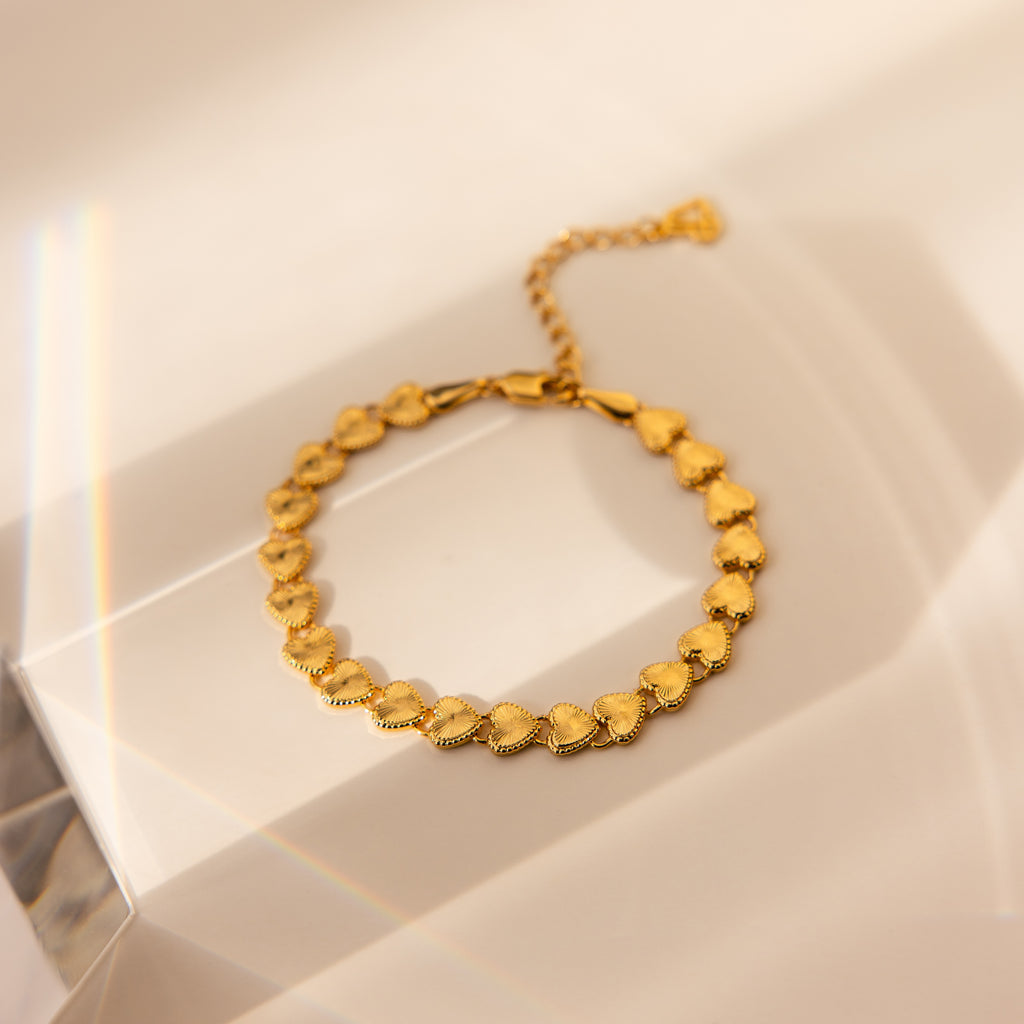 A delicate gold bracelet is artfully displayed on a soft, cream-toned geometric surface. The bracelet is composed of a continuous series of ribbed heart-shaped links, each with fine sunburst-like detailing that radiates from the center of each heart.  The hearts are evenly spaced and connected seamlessly
