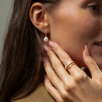 A woman touches her face, showcasing manicured nude nails and Pearl Diamond Drop Earrings in 18K Gold.