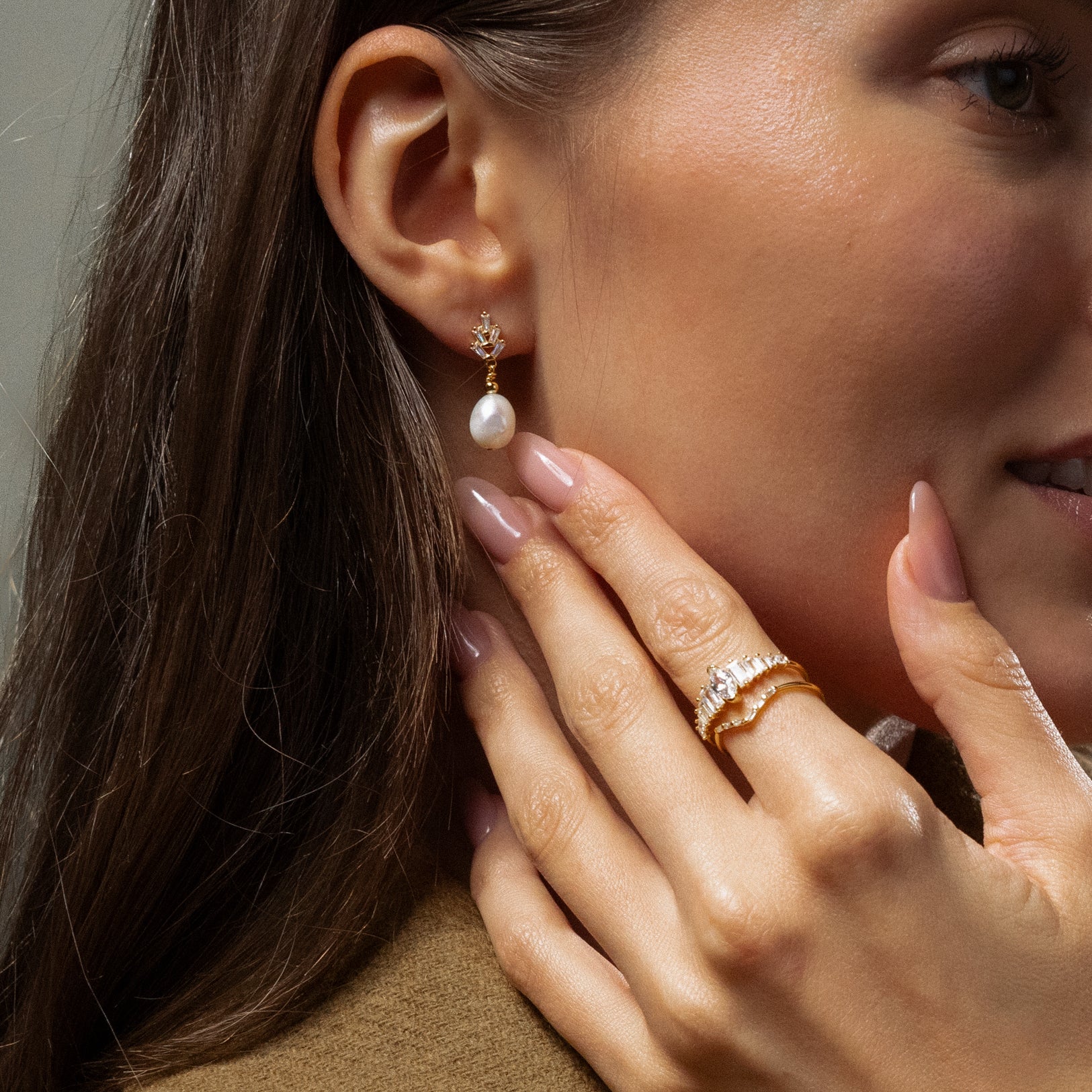 A woman touches her face, showcasing manicured nude nails and Pearl Diamond Drop Earrings in 18K Gold.
