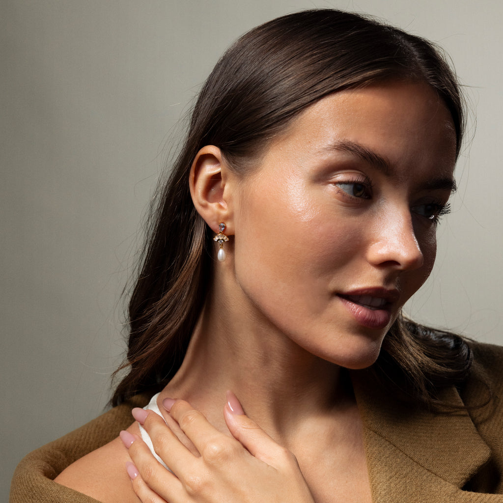 Woman with long brown hair in a brown blazer wears Aquamarine Pearl Drop Earrings in 18K Gold, exuding vintage glamour as she gazes thoughtfully to the side.