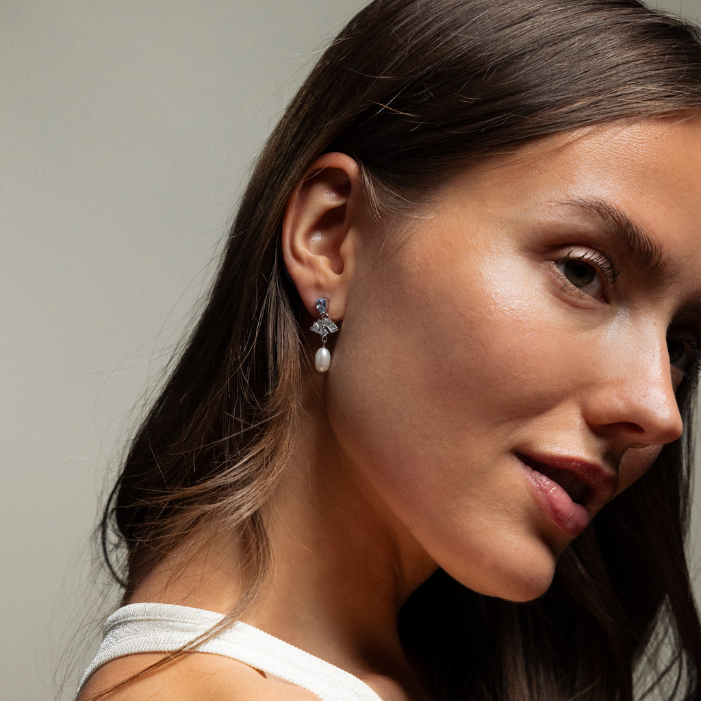 Woman with long brown hair, in a white top and Aquamarine Pearl Drop Earrings in Silver, gazes softly at the camera, adding a touch of vintage glamour.