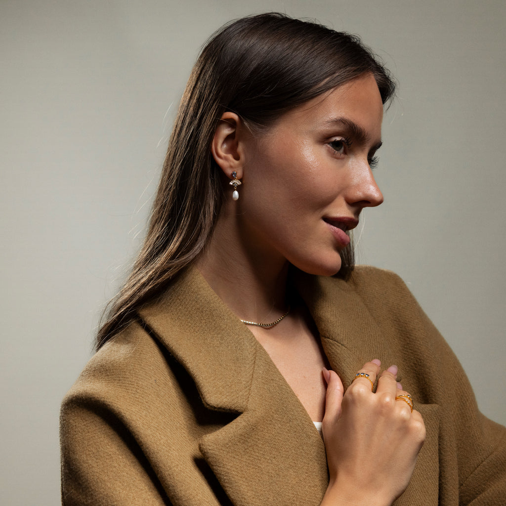 Woman in a brown coat wears Aquamarine Pearl Drop Earrings in 18K Gold, along with a gold necklace and rings, radiating vintage glamour as she gazes to the side.