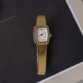 The Petite Mesh Timepiece features a gold rectangular face with diamond-like stones around the dial, shown elegantly on a dark surface.