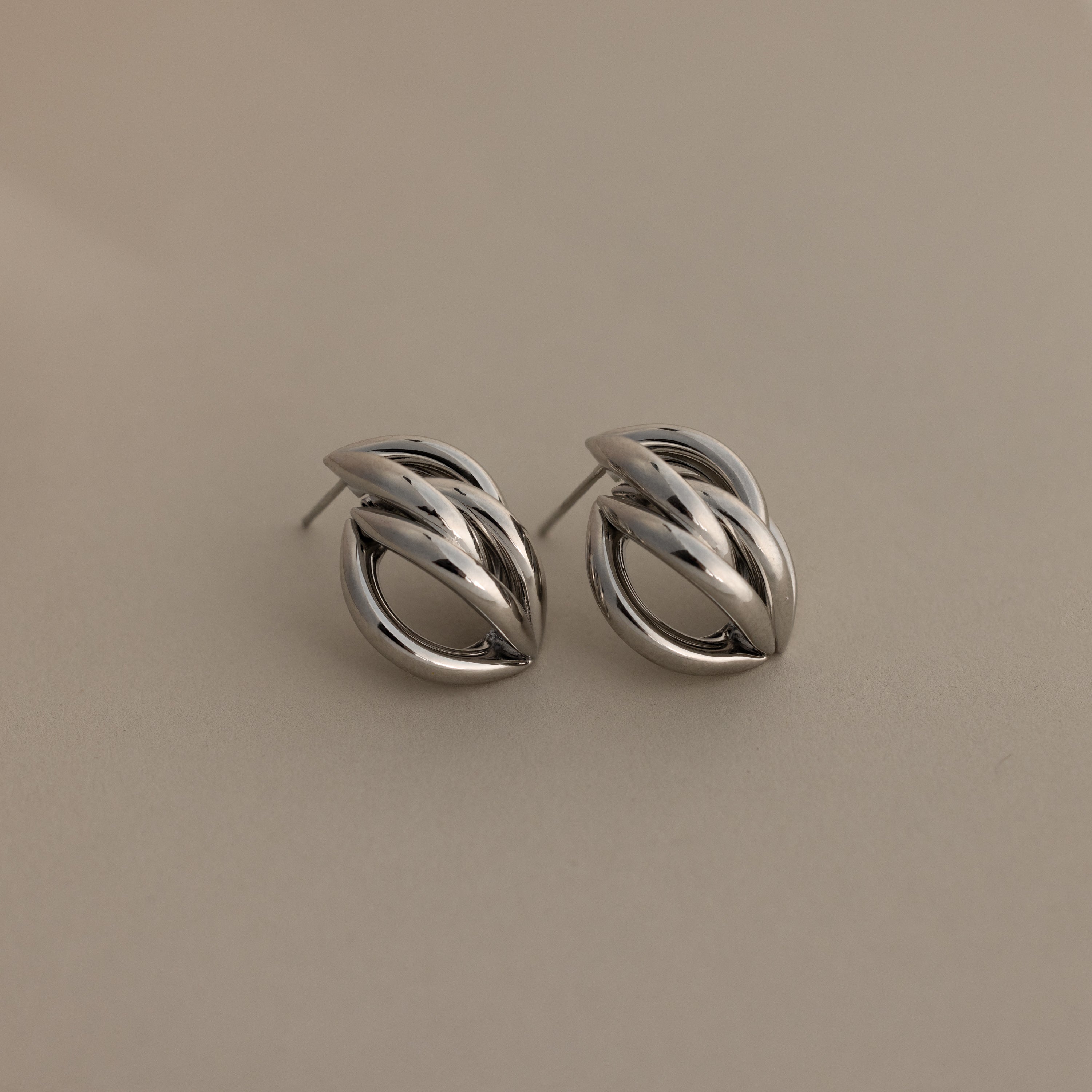 The Bold Multi Knot Studs in silver feature an intricate knot design, embodying modern elegance against a light beige background.