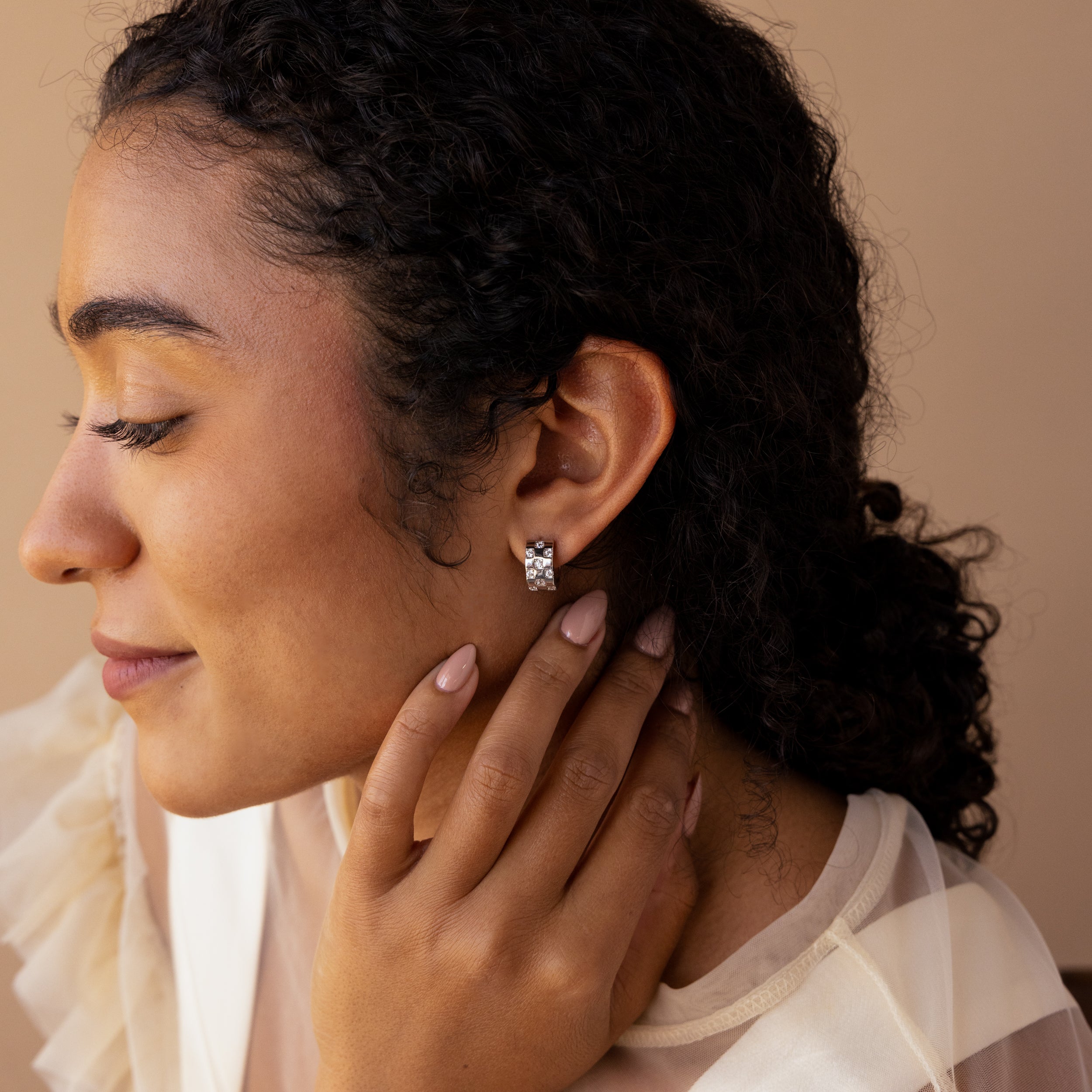 Woman with curly hair in a sheer cream blouse and silver floral earrings, showcasing the Pave Checkered Huggies in Silver as chunky hoops for a modern touch.