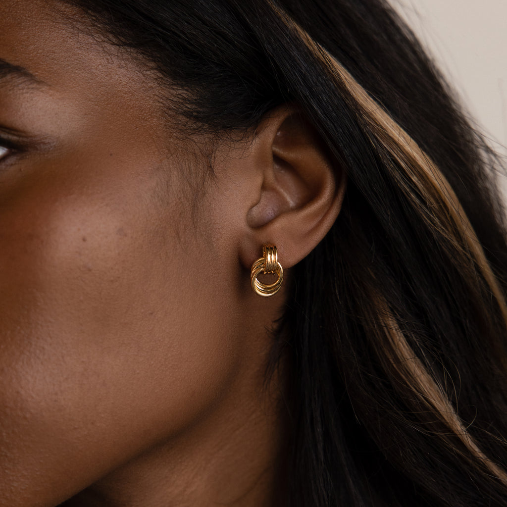 Close-up of a womans ear with dark hair and a light streak, wearing Micro Triple Knot Earrings in 18K Gold, embodying quiet luxury.
