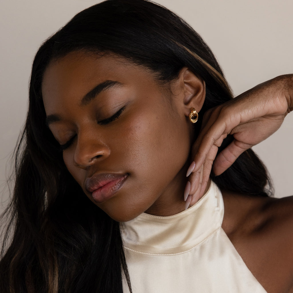 Woman with long dark hair and eyes closed, posing with hand near face, wearing Micro Triple Knot Earrings in 18K Gold—evoking a sense of quiet luxury.