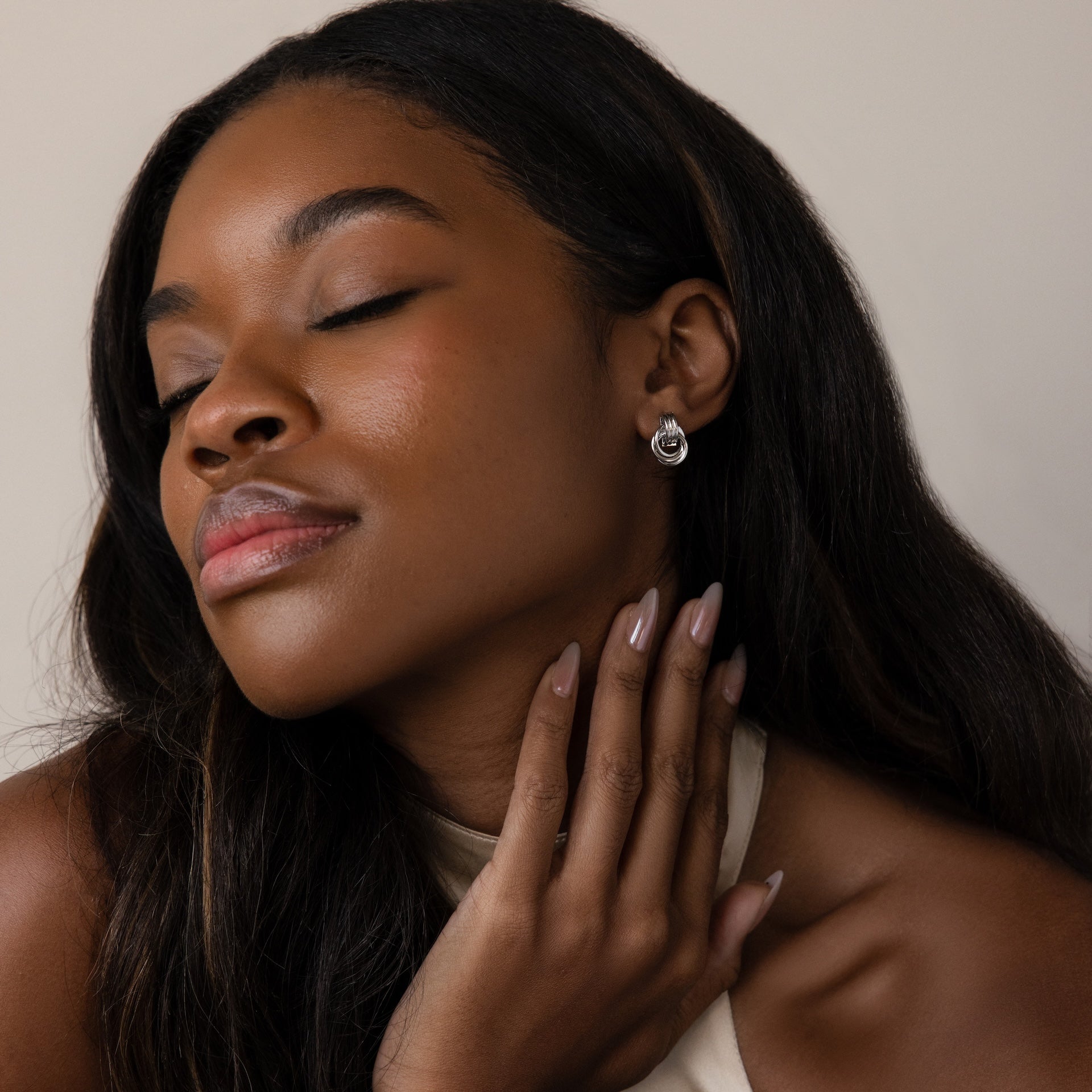 Woman with long dark hair and closed eyes touches her neck, wearing Micro Triple Knot Earrings in Silver and a cream top.