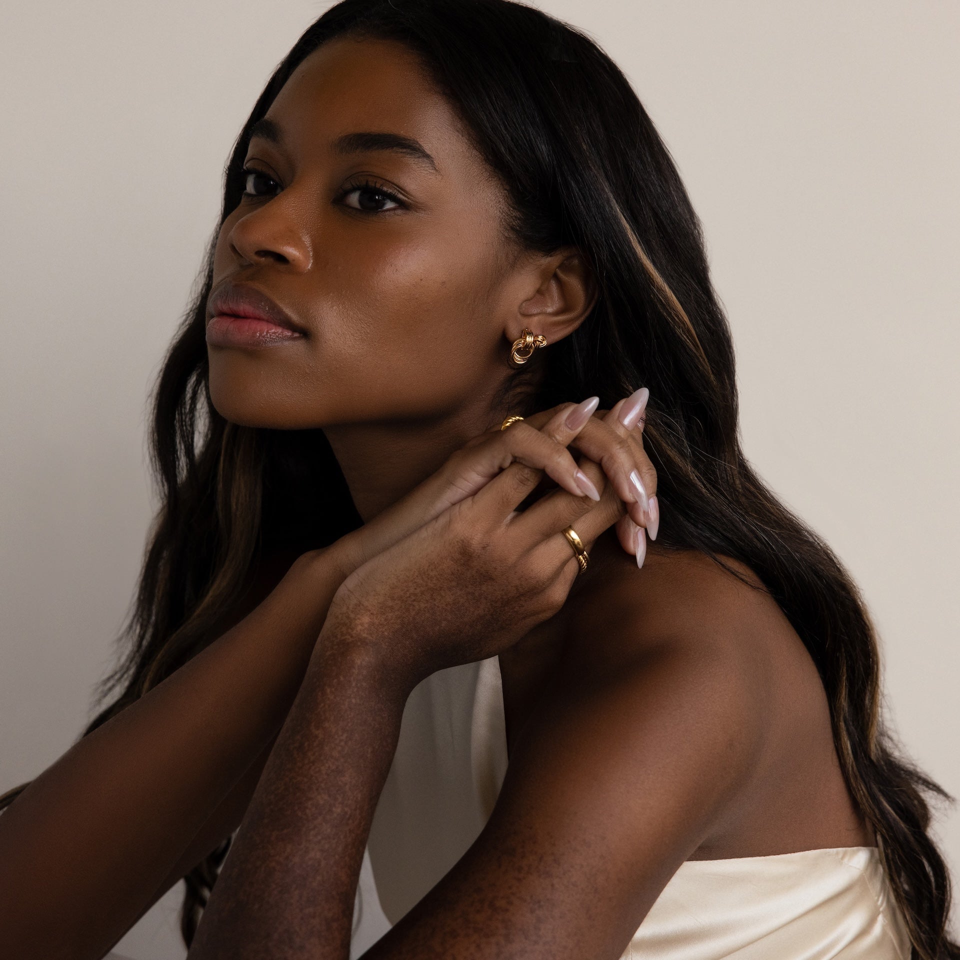 Woman with long dark hair wearing Micro Triple Knot Earrings in 18K Gold and a cream top, posing with hands near her face for a look of quiet luxury against a plain background.