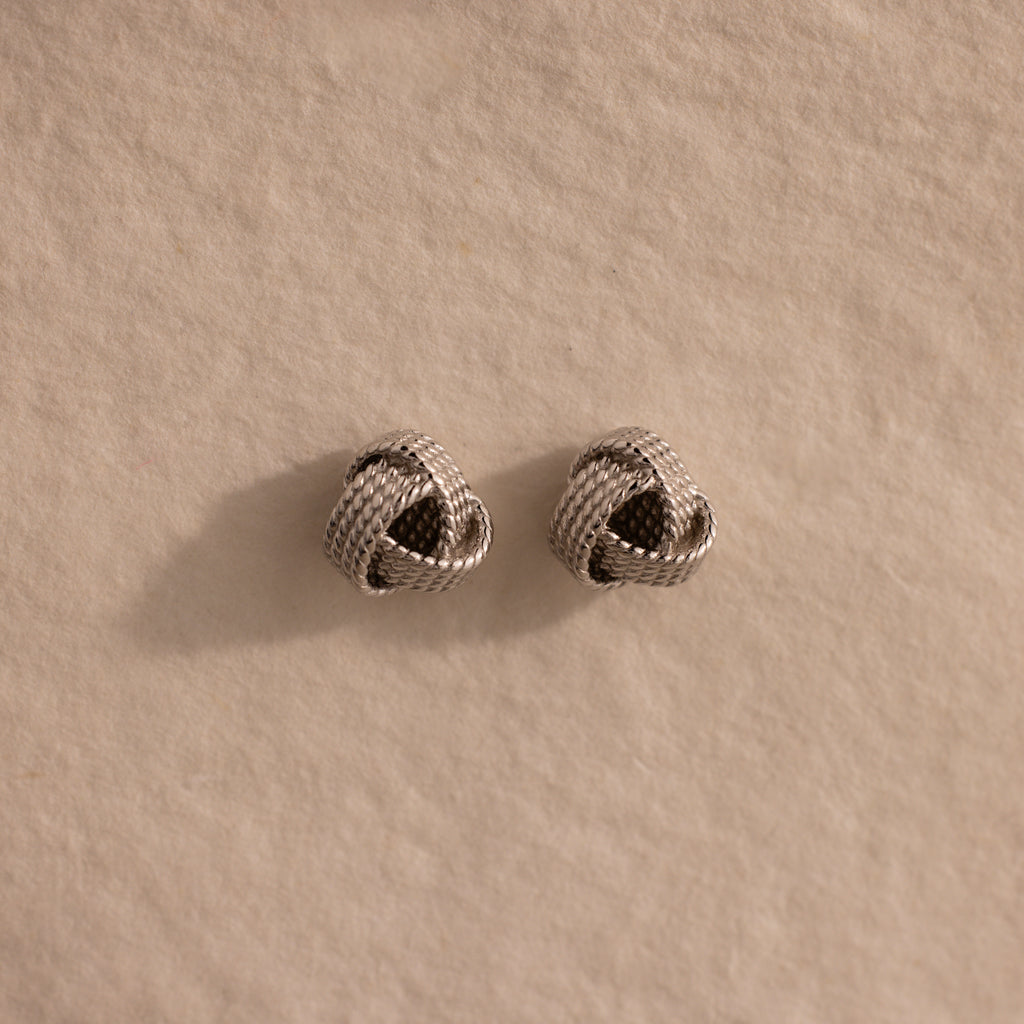The Rope Knot Studs in Silver, with their elegant knot design, rest on a textured beige surface—timeless accessories that are perfect for any jewelry collection.