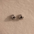 The Rope Knot Studs in Silver, with their elegant knot design, rest on a textured beige surface—timeless accessories that are perfect for any jewelry collection.