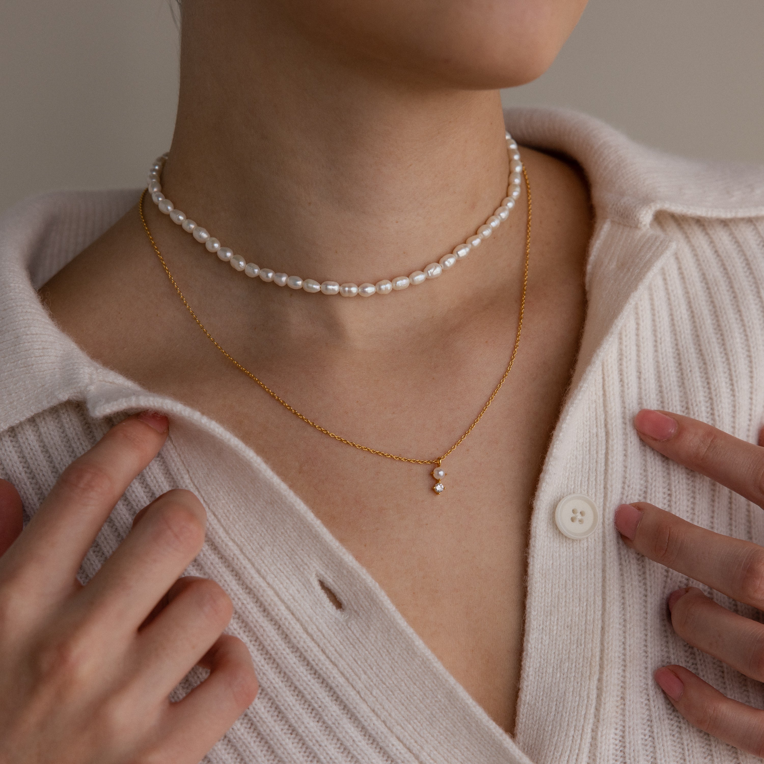 A person wears a pearl choker and the Pearl Diamond Drop Necklace in 18K Gold over a cream ribbed cardigan.