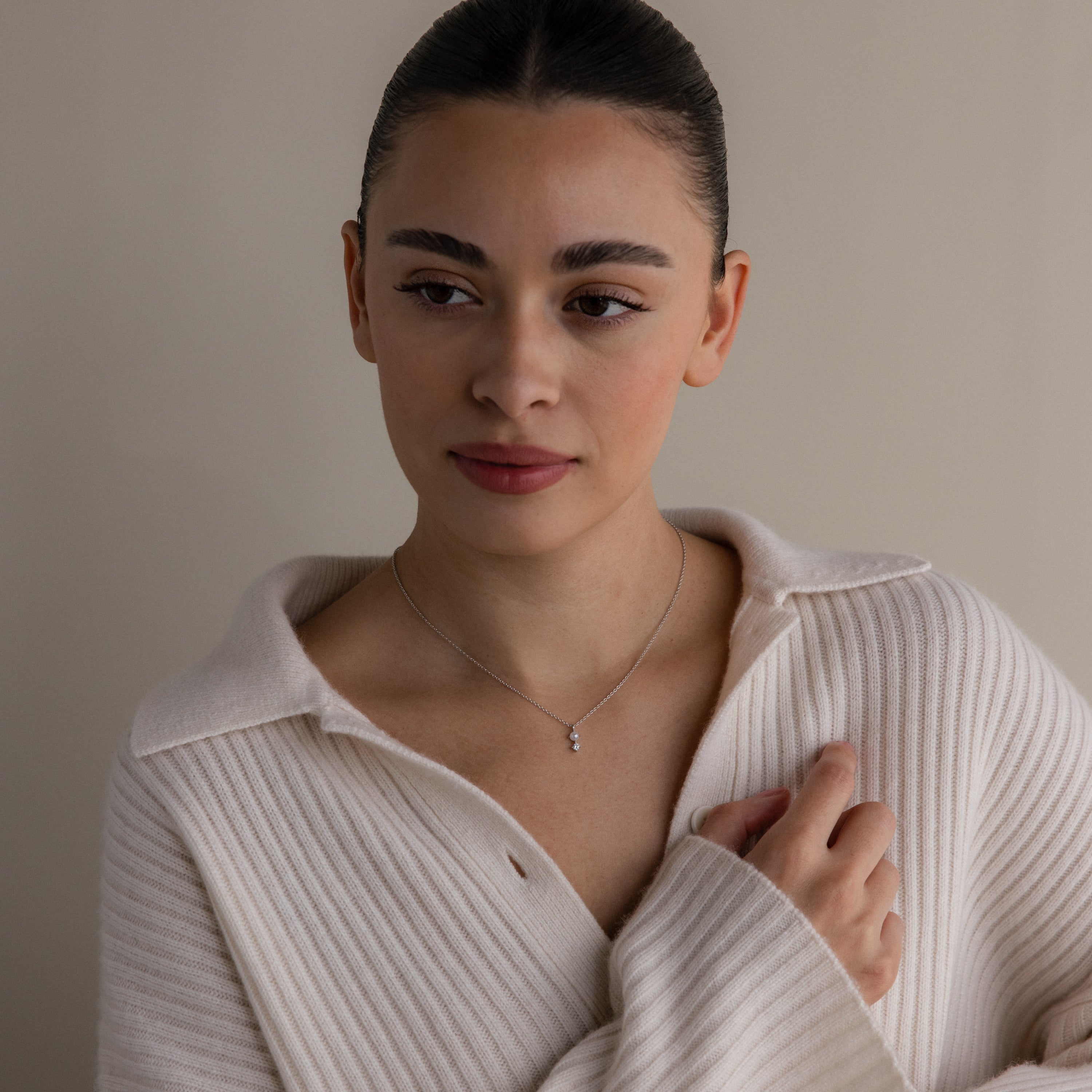 Woman in a cream ribbed sweater wearing the Pearl Diamond Drop Necklace in Silver, looking to the side against a neutral background.