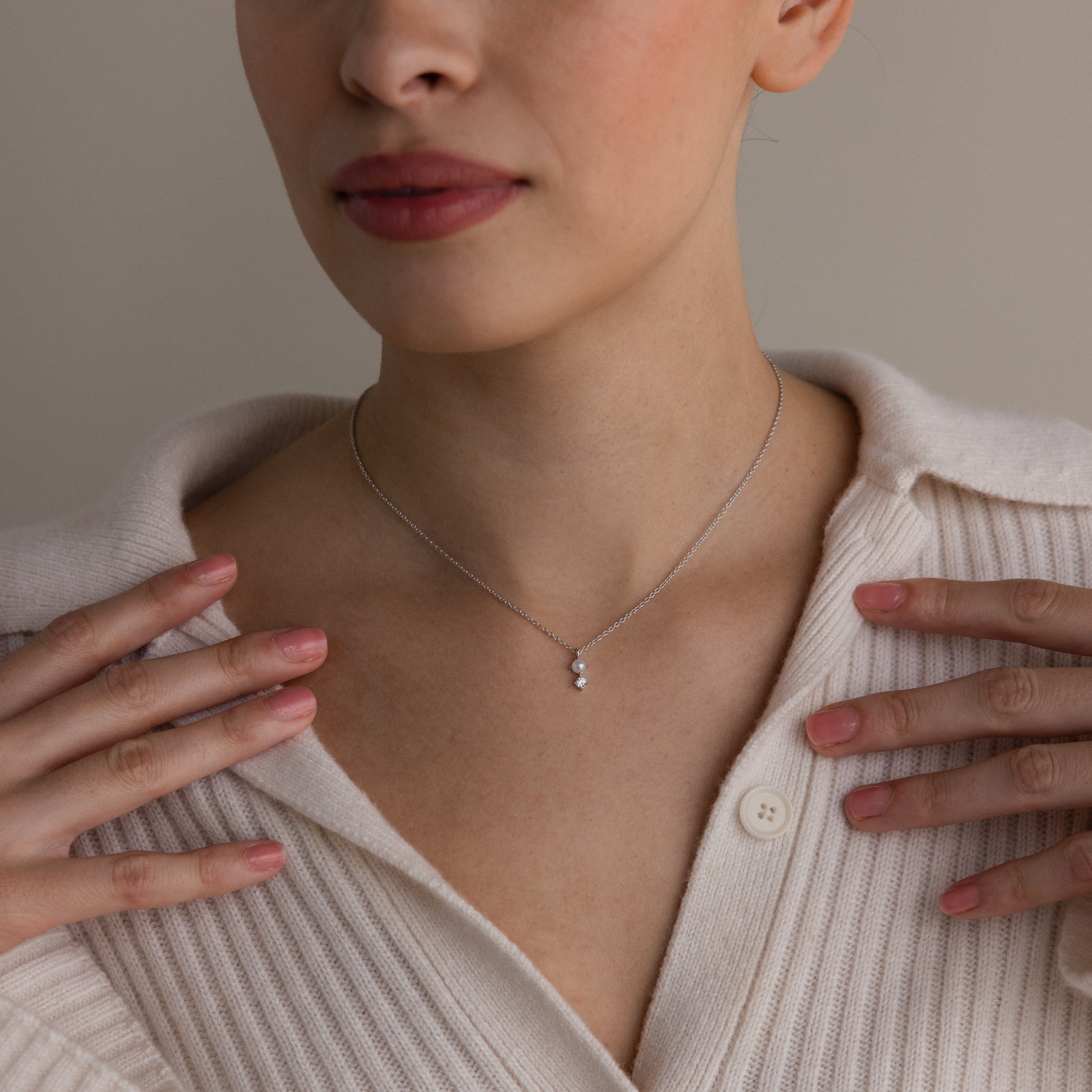 A woman wearing a cream sweater gently touches her collarbone, showcasing the Pearl Diamond Drop Necklace in Silver with its delicate pendant.