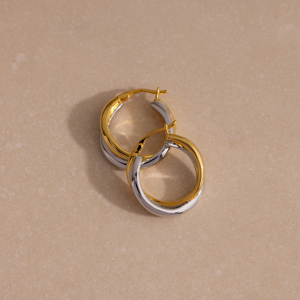 The Twisted Mixed Metal Hoops in gold and silver are displayed on a beige, lightly textured surface, highlighting their modern chic design.