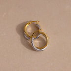 The Twisted Mixed Metal Hoops in gold and silver are displayed on a beige, lightly textured surface, highlighting their modern chic design.