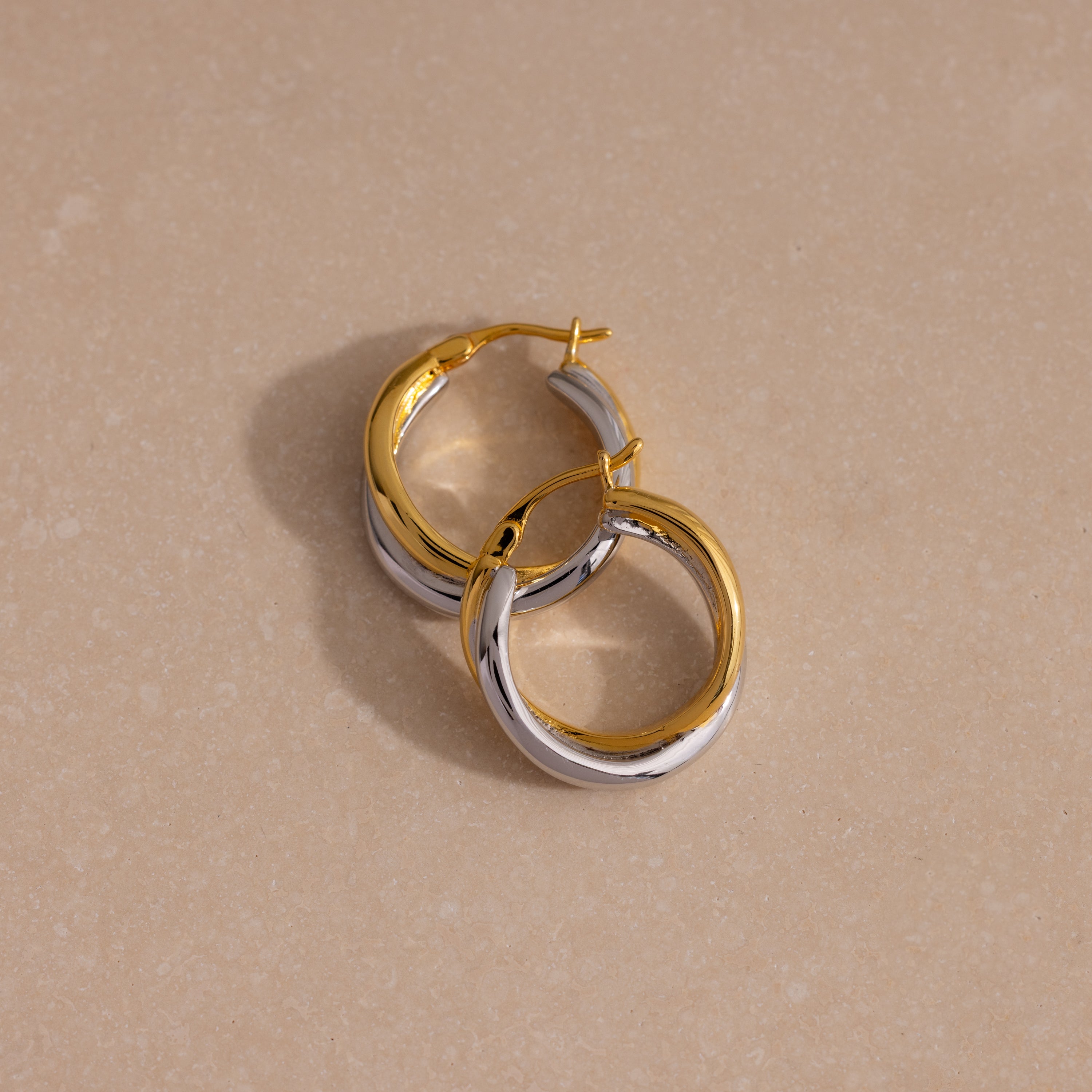 The Twisted Mixed Metal Hoops in gold and silver are displayed on a beige, lightly textured surface, highlighting their modern chic design.