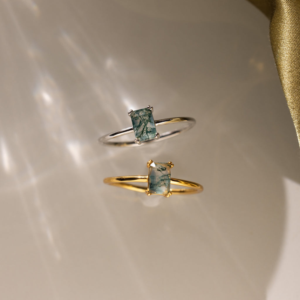 A side-by-side view of the Agate Emerald Ring in both gold and silver finishes on a glossy surface with light reflections.