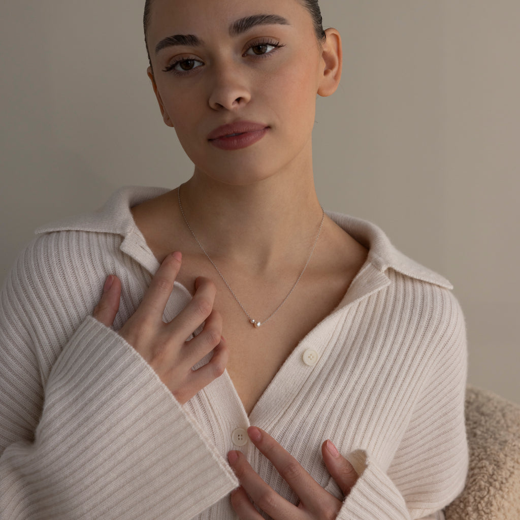 Woman in a cream ribbed sweater gently touches the Dainty Duo Pearl Necklace, looking calmly at the camera—an elegant choice for wedding jewelry.