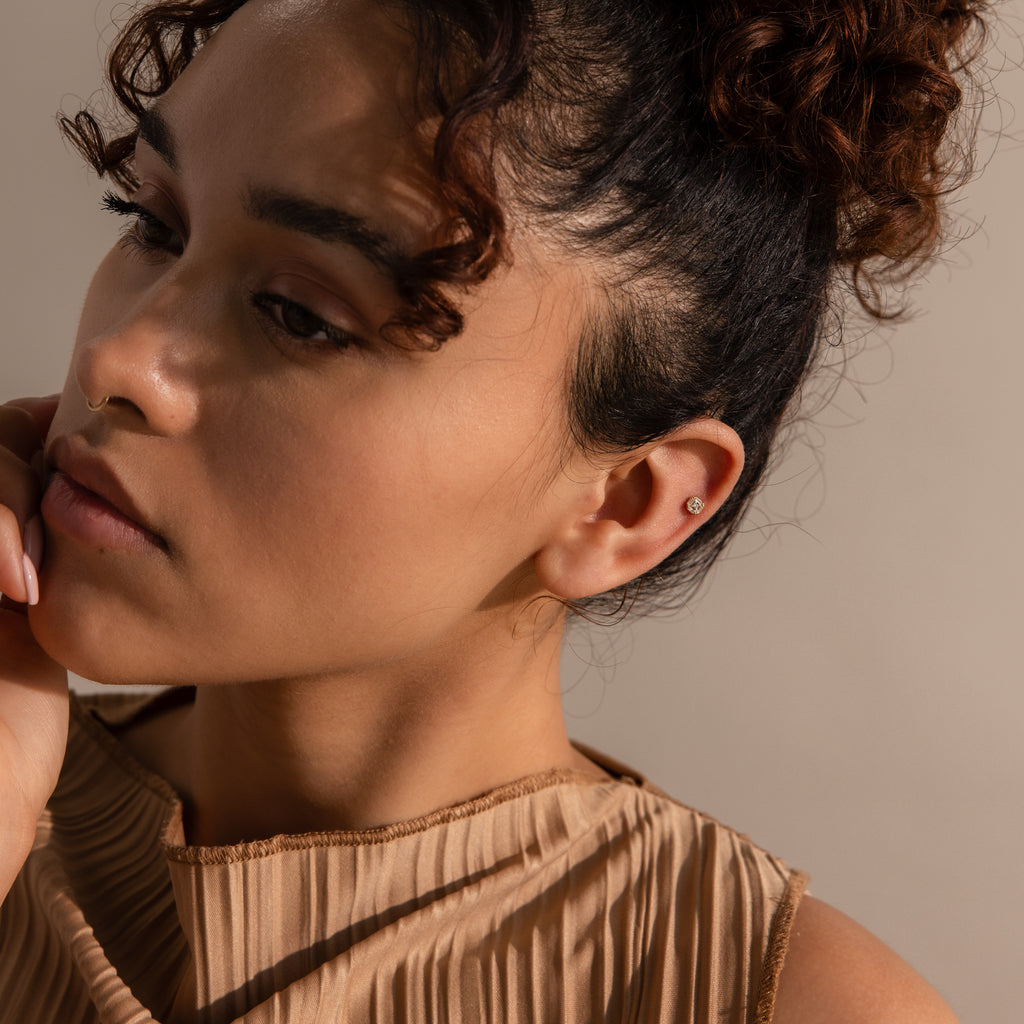 Model with curly hair wearing a tiny square diamond stud in her upper ear, gazing downward.